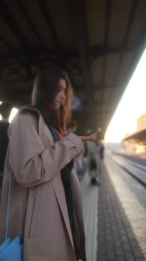 Genç bir kadın gün batımında bir tren istasyonunda sabırla beklerken akıllı telefonuna dalmış bekliyor.