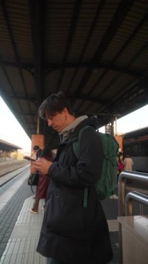 Genç bir adam hareketli bir tren istasyonunda duruyor, diğer yolcuların arasında akıllı telefon cihazına kendini kaptırmış.