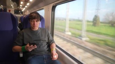 Genç bir adam bir tren kulübesinde dinleniyor, akıllı telefonuna odaklanıyor ve dışarıdaki manzaranın tadını çıkarıyor.
