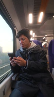 Genç bir birey, trende giderken akıllı telefonlarına dalmış, modern banliyö uygulamalarını yansıtıyordu.