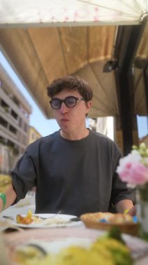 Genç bir adam açık bir kafede yemek ve içkiden zevk alıyor, rahat bir yemek atmosferi yakalıyor.