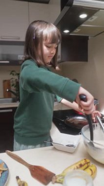 Genç bir kız neşeyle krep pişirir, yeteneklerini renkli mutfak malzemeleriyle dolu parlak bir mutfakta sergiler.