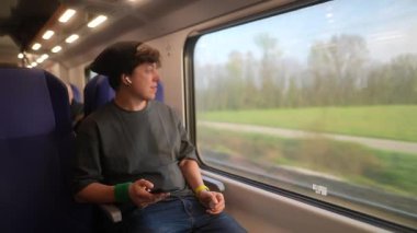 Genç bir insan akıllı telefonuna dalmış, trende dijital içeriğin tadını çıkarırken manzarayı seyrediyor.