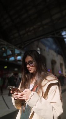 Modayı takip eden genç bir kadın telefonuna birçok yolcuyla çevrili, hareketli bir tren istasyonunda bakar.