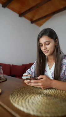 Rahat bir bornoz giymiş genç bir kadın şık ve modern bir yaşam alanında akıllı telefonuyla etkileşime giriyor.