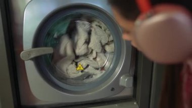 Genç bir insan modern bir makinede yıkanan kıyafetleri dikkatle izler, günlük çamaşırların rutinini gösterir.
