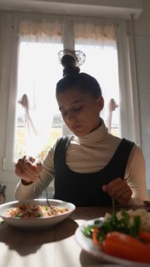 Genç bir kadın güneşli bir mutfak masasında besleyici bir yemekten zevk alıyor, canlı sebzeler sergiliyor..