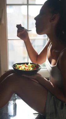 Bir kadın mutfak tezgahında canlı bir salata yemekten hoşlanır. Pencereden gelen sıcak doğal ışıkla aydınlatılır.