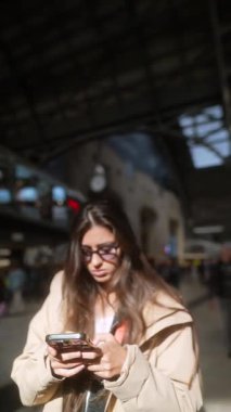 Aktif olarak akıllı telefonuyla meşgul olan genç bir kadın bir tren istasyonunun telaş ve telaşıyla çevriliyken...