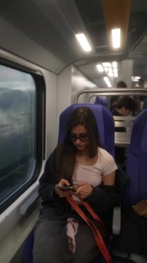 Şık güneş gözlüğü takan genç bir kadın trende rahat bir şekilde seyahat ederken akıllı telefonuyla derinden meşgul olur.