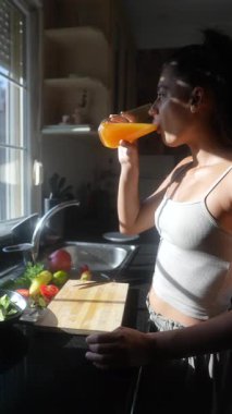 Neşeli bir insan güneşli bir mutfakta bir bardak meyve suyu içerken neşeyle taze sebze hazırlar.