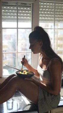 Bir kadın mutfak tezgahına oturmuş, bir kase salatanın tadını çıkarıyor, pencereden güneş ışığı akıyor..