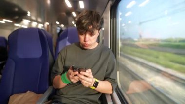 Genç bir gezgin, trende seyahat ederken dikkatle akıllı telefonuna odaklanır ve dışarıdaki manzaranın tadını çıkarır.