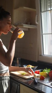 Bir kadın, güneşli ve sıcak mutfağında içkisini yudumlarken bir dizi taze sebzeyi ustalıkla hazırlar.