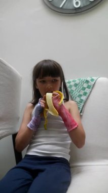 Parlak, renkli bir bluz giymiş genç bir çocuk sıcak ve neşeli bir ortamda muz yemekten mutlu olur.