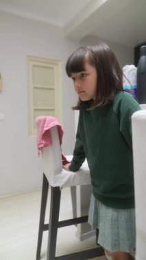 Parlak yeşil bir kazak giyen genç bir çocuk düşünceli bir şekilde bir sandalyenin yanında dururken oldukça dalgın görünür.