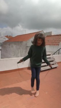 Genç bir kız neşeyle çatıda ip atlıyor, çevikliğini ve neşesini bulutlu bir gökyüzünün altında sergiliyor..