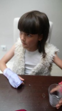 Renkli bandajlarla süslenmiş genç bir kız düşünceli bir şekilde bir masaya oturur, derin derin düşüncelere dalmış bir bardağa bakar.