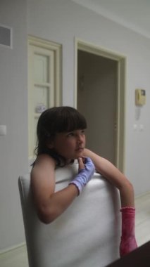 Genç bir kız güzel düzenlenmiş ve dekore edilmiş bir masada oturuyor, renkli eldivenler giyiyor, düşüncelerinde derinlere dalmış.