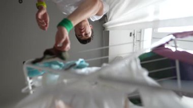 Genç bir insan, parlak ve neşeli bir iç mekanda kurutma askısına çeşitli giysiler asıyor.