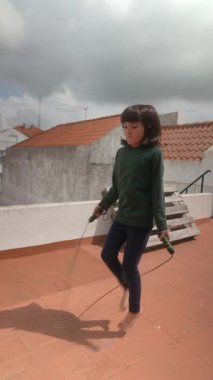 Mutlu bir çocuk neşeyle çatıda ip atlıyor, eğlenceli ve sağlıklı açık hava oyun deneyimlerini teşvik ediyor.