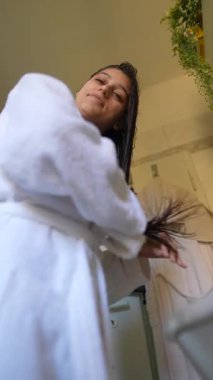 Sıcak bornoz giymiş bir kadın, sakin ve rahatlatıcı bir banyo ortamında saçını tararken mutlu bir şekilde görülüyor.