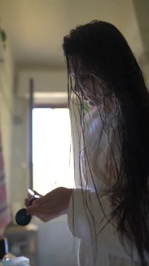 Dalgalı beyaz bornoz giymiş bir kadın, güzellik ürünlerini parlak, havadar bir banyoya uyguluyor.