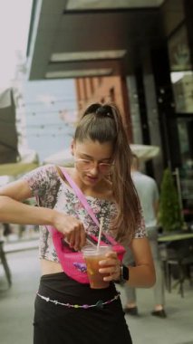 Şık, genç bir kadın, canlı bir şehir ortamında serinletici bir içkiden zevk alır, gündelik ama şık tarzını sergiler.