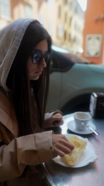 Tarz sahibi genç bir kadın rahat bir kafede oturmuş kahveyle eşleştirilmiş lezzetli bir pastanın tadını çıkarıyor.