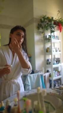 Bir kadın cilt ürününü cazibeyle dolu parlak ve iyi korunmuş bir banyoya uyguluyor.