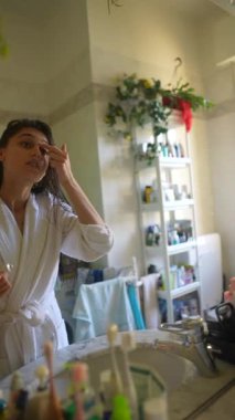 Parlak, güneşli bir banyoda, rahat bir bornoz giymiş bir kadın sabah cilt bakımını titizlikle yapar.