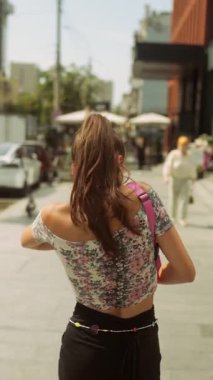 Çiçeklerle süslenmiş şık bir genç kadın canlı bir şehir caddesinde geziyor, şehir modasını gözler önüne seriyor..