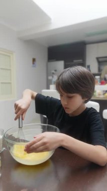 Neşeyle dolu genç bir çocuk, parlak, güneşli bir mutfakta yumurta çırpıyor, yaratıcılık ve aşçılık keyfi sergiliyor.