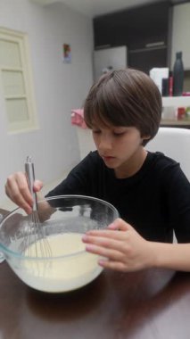 Parlak ve modern bir mutfak ortamında, odaklanmış ve kararlı bir çocuk hamuru dikkatlice çırpıcıyla harmanlıyor.