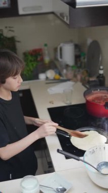 Genç bir çocuk mutfakta neşeyle yemek pişiriyor, mutlu bir şekilde krep hazırlıyor ve tüm sürecin tadını çıkarıyor.