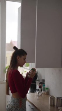 Parlak bir mutfakta, genç bir kadın taze yeşil bir meyveyi denetler, sağlık ve yaşam tarzı seçimlerini sembolize eder.