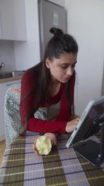 Genç bir kadın, şık bir mutfak ortamında rastgele elma yerken tabletine bir görev yüklemiş..
