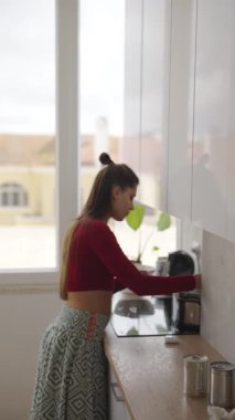 Bir kadın, doğal ışık ve canlı bitkilerle dolu aydınlık, modern bir mutfakta heyecanlı bir şekilde yemek pişiriyor.