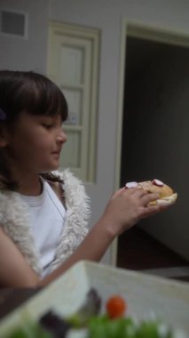 Genç bir kız, yemek masasında oturmuş, keyifli bir anı yakalayan bir sandviç tutuyor..