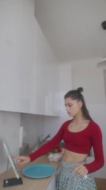 Genç bir kadın modern bir mutfakta zevkle lezzetli bir yemek pişirir. Yeteneklerini göstermek için dijital bir tarif kullanır.