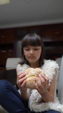 Genç bir kız evde rahat bir şekilde oturmuş, elinde bir somun taze ekmeği tutarken dikkatlice tadını çıkarıyor.