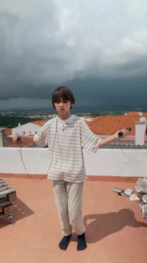 Genç bir çocuk neşeyle çatıda ip atlıyor, dramatik bir bulutla çevrili, gençlik ruhunu sergiliyor.