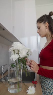 Bir kadın ustalıkla ve zarafetle mutfak tezgahındaki cam vazoda narin beyaz çiçekler düzenler.