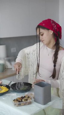 Havalı bir mutfakta şık genç bir kadın parlak kırmızı bir bandanayla yumurta çırpıyor.