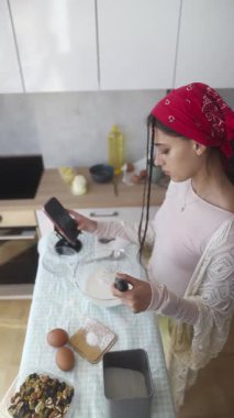 Bir kadın, şık bir mutfakta akıllı telefonunu kontrol ederken aynı anda çeşitli bileşenleri doğru bir şekilde ölçer.