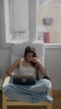 Genç bir kadın rahat bir şekilde bir sandalyeye oturur, dizüstü bilgisayarına odaklanır, evinde sıcak ışığın tadını çıkarır.