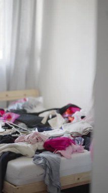Dağınık bir şekilde yatağın her tarafına dağılmış renkli ve canlı giysiler sergilenen dağınık bir yatak odası sahnesi.