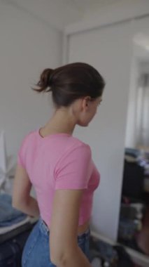Genç bir kadın dağınık yatak odasında çeşitli şık giysiler deniyor, oyuncu moda anlayışını sergiliyor.