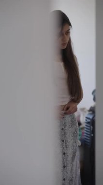 Genç bir insan farklı giysiler dener. Güzelce tasarlanmış, biraz dağınık bir odada doğal ışık altında.