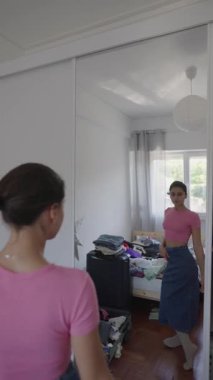 Modayı takip eden genç bir kadın şık odasında modellik yapıyor. Kıyafet seçimlerini ve ayna yansımasını gösteriyor.